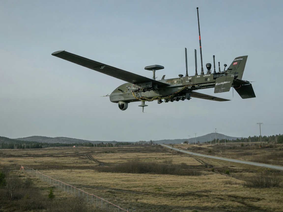Militær drone i flukt over åpent landskap, med antenner og kommunikasjonsutstyr synlig på skroget.