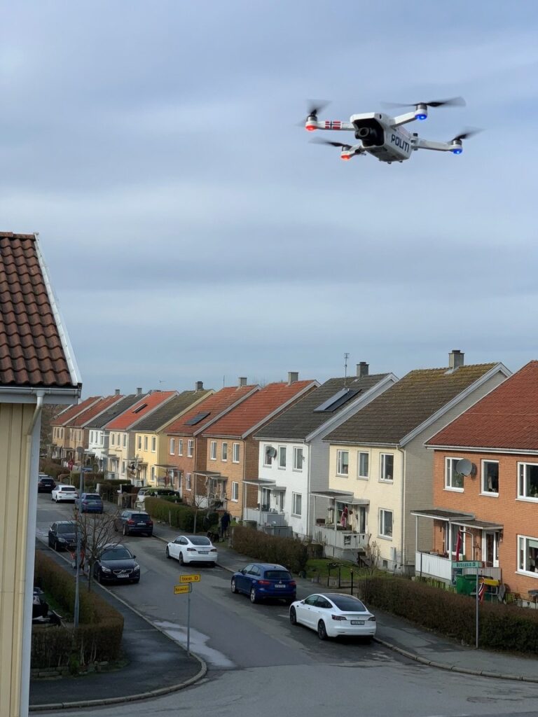 Liten politidrone i luften over et boligstrøk, med urbant landskap i bakgrunnen