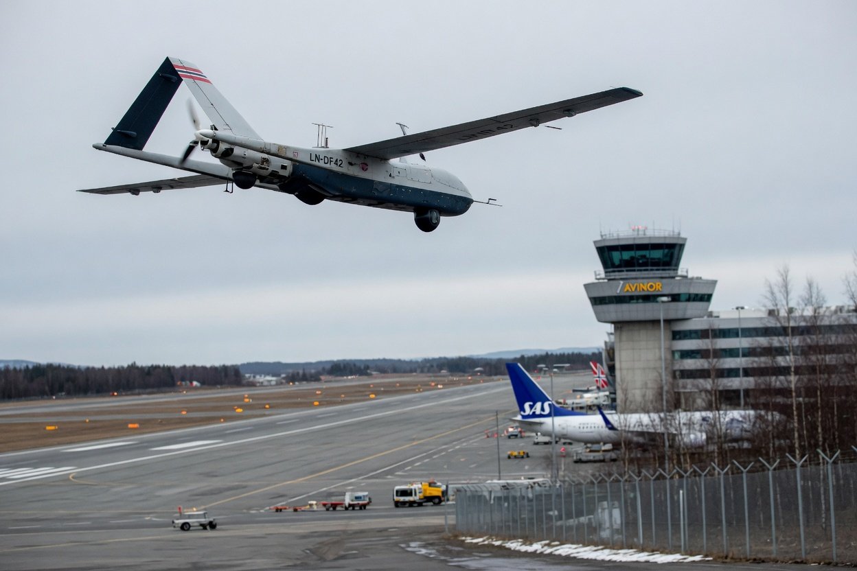 Stor fastvinget drone i luften over flyplassmiljø med kontrolltårn i bakgrunnen