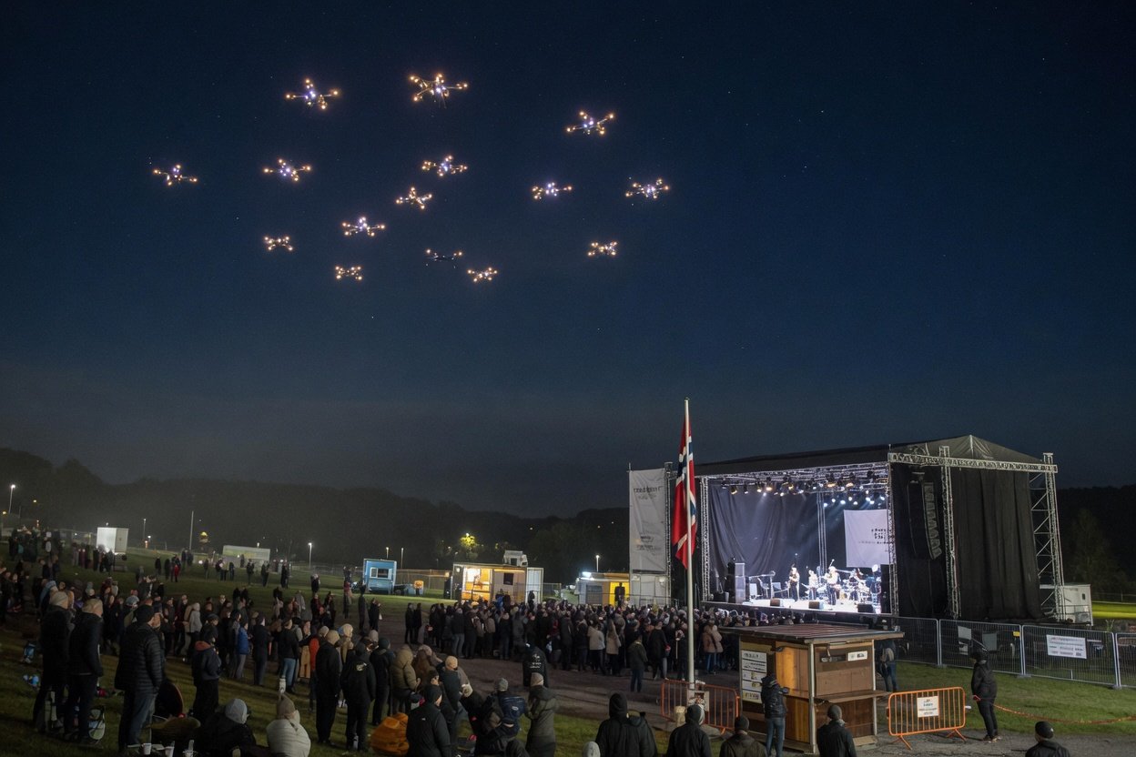 Lysende droneformasjon i mørket over stor utendørs konsertscene med publikumsområde nedenfor