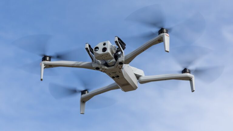 Skydio X10 i luften mot blå himmel, sett nedenfra med kamerahodet synlig