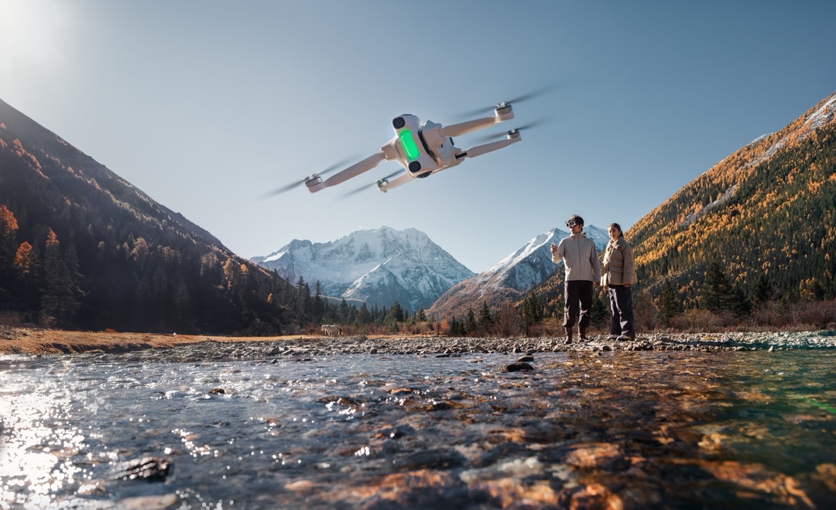 Antigravity A1-drone i lufta over en fjellelv, med to personer og snødekte fjell i bakgrunnen