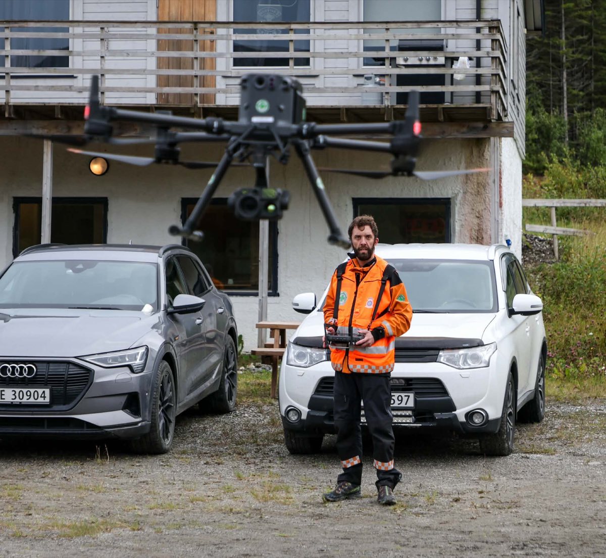 Pilot i oransje redningsjakke med fjernkontroll, mens en stor quadkopterdrone svever foran ham