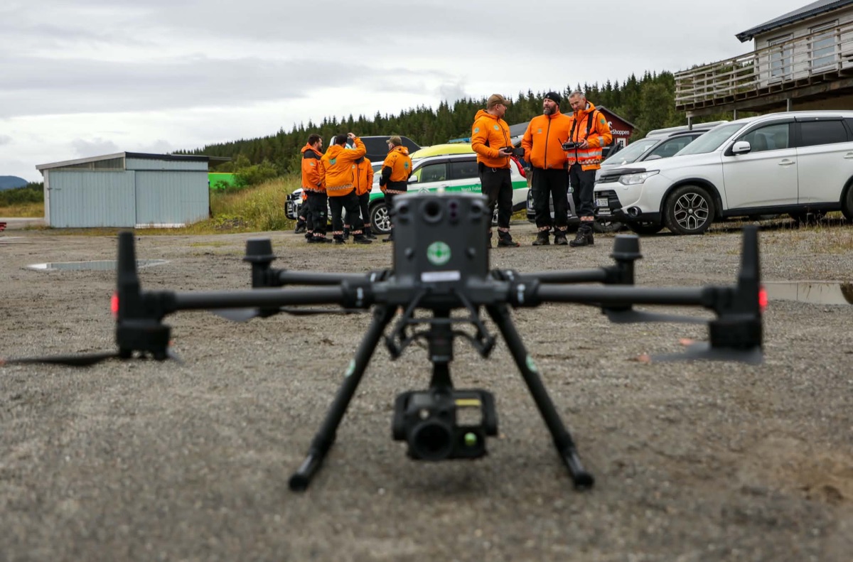 Industridrone på grusplass i forgrunnen, med mannskaper i oransje redningsjakker og biler i bakgrunnen