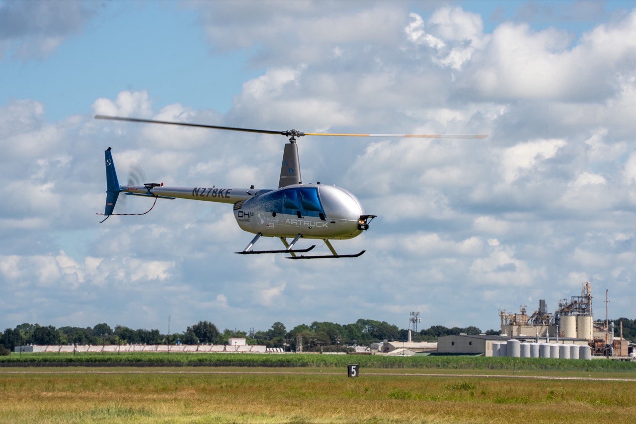 Robinson Unmanned R44 AIRTRUCK, et fjernstyrt lastehelikopter uten cockpitvinduer, svever lavt over en flyplass med industrianlegg i bakgrunnen