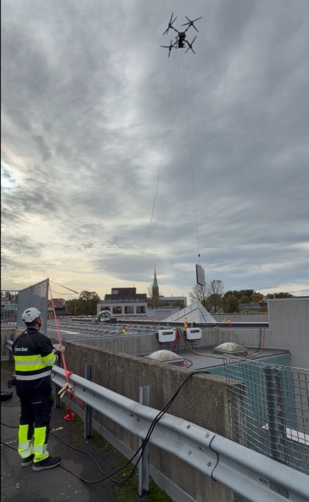 En DJI FlyCart 100 løfter et panel i line opp mot et tak der en mann i vernetøy styrer en hjelpeline