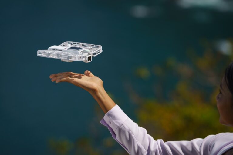 Den hvite flyvende kameradronen HoverAir VERSA svever over en utstrakt hånd med havet i bakgrunnen
