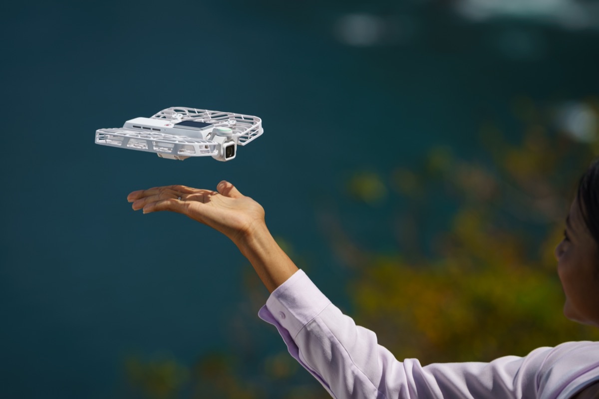 Den hvite flyvende kameradronen HoverAir VERSA svever over en utstrakt hånd med havet i bakgrunnen