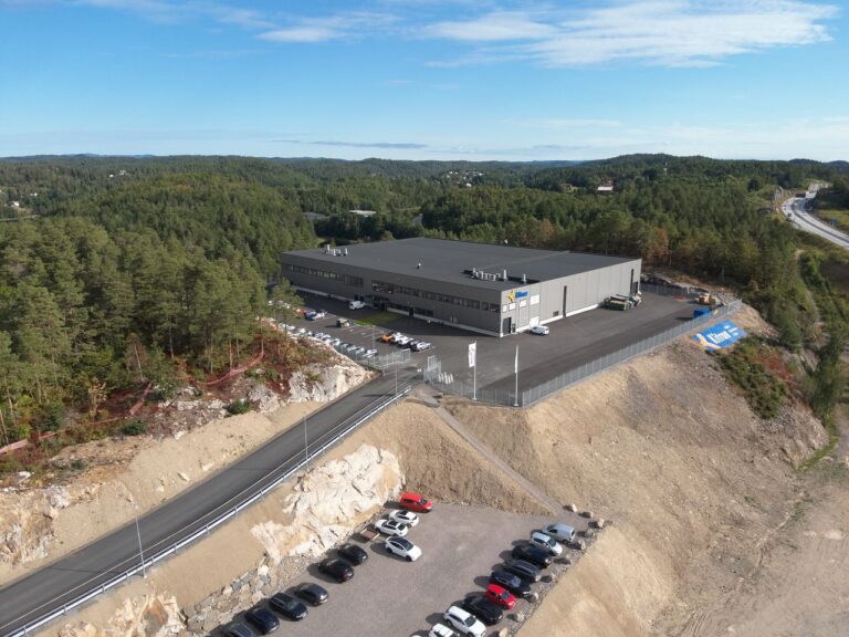 Dronefoto av Kitrons nye fabrikkbygg i Arendal, med parkeringsplass, gjerde og skog rundt